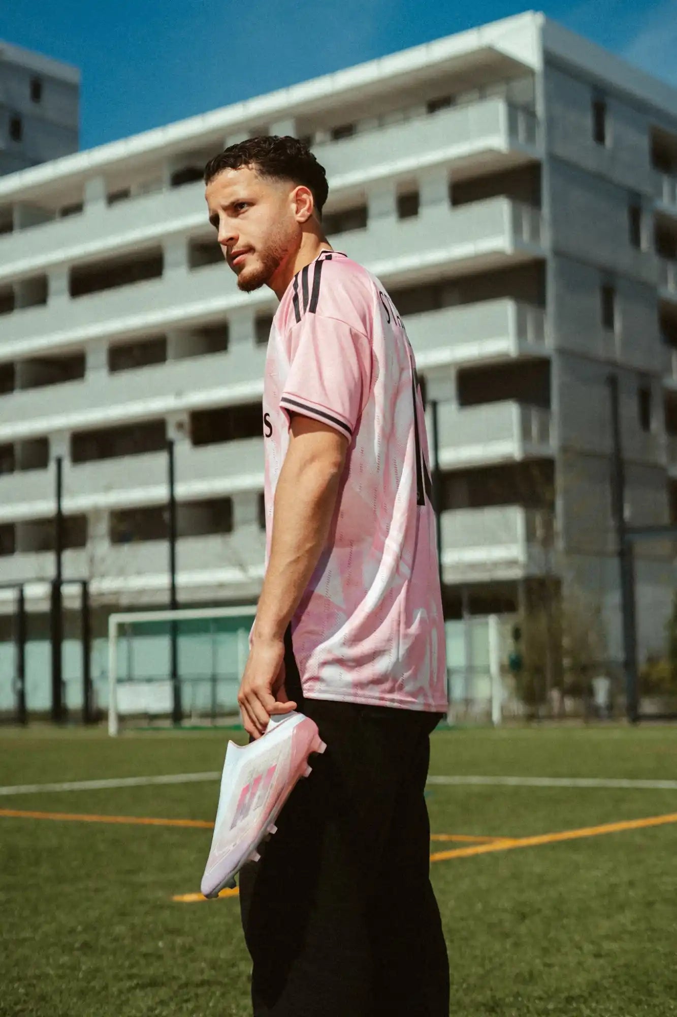 Un homme en chemise de sport rose et pantalon noir tenant une paire de crampons de football blancs et roses.
