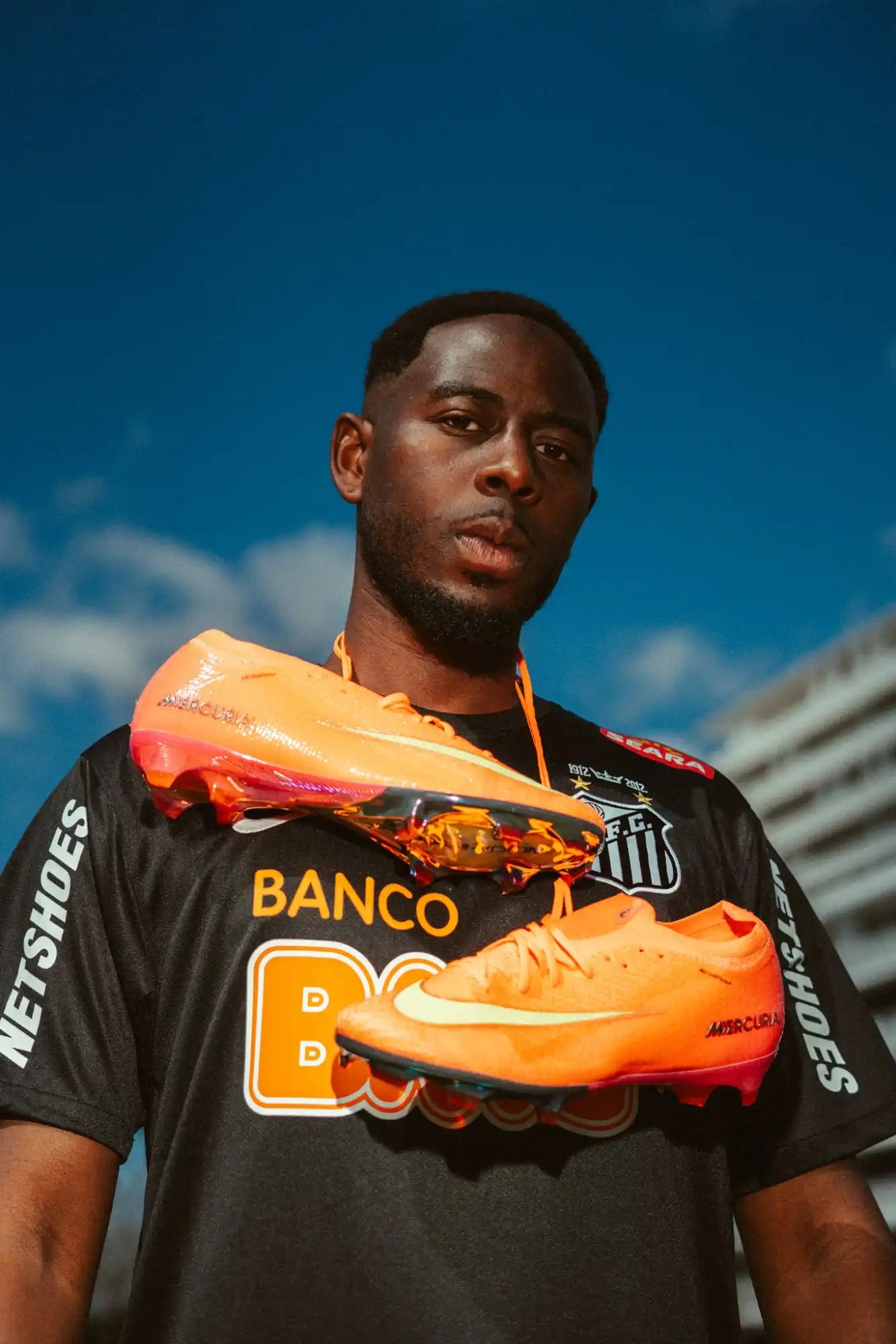 Un homme à la peau foncée portant une chemise noire avec un motif blanc et orange, tenant une paire de crampons de football orange vif et rouge.